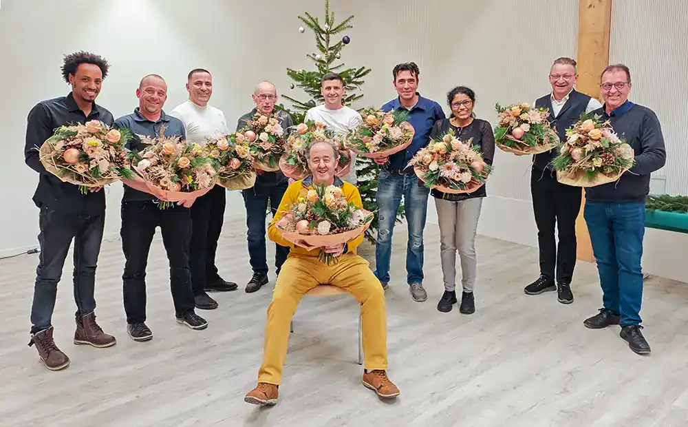 Un groupe de personnes au sourire amical, debout devant un sapin de Noël décoré et tenant des arrangements floraux