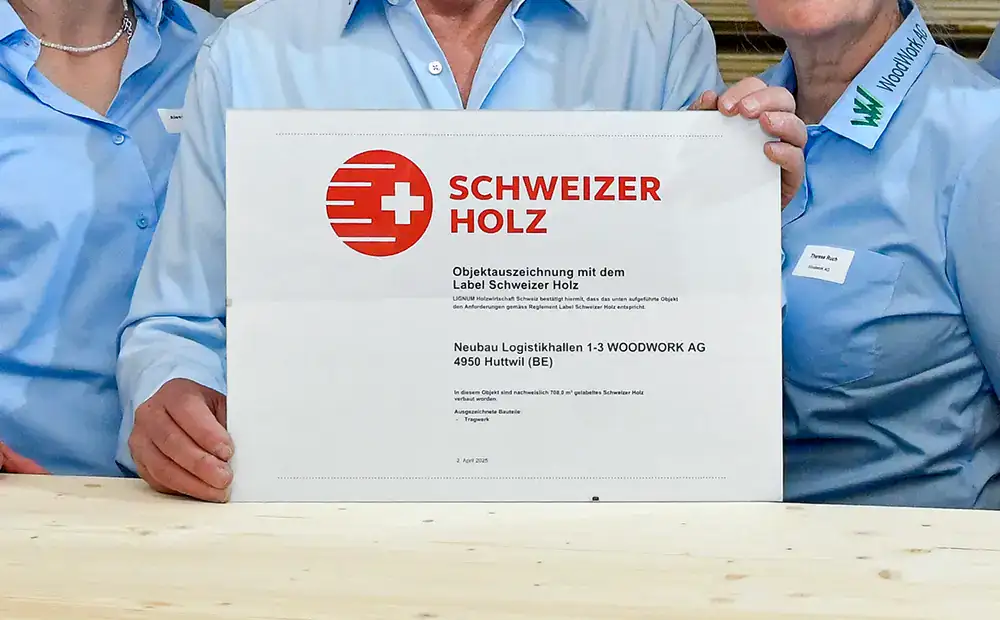 EIn Händepaar von Menschen in hellblauen Firmenhemden hält ein gerahmtes Zertifikat mit Text Schweizer Holz